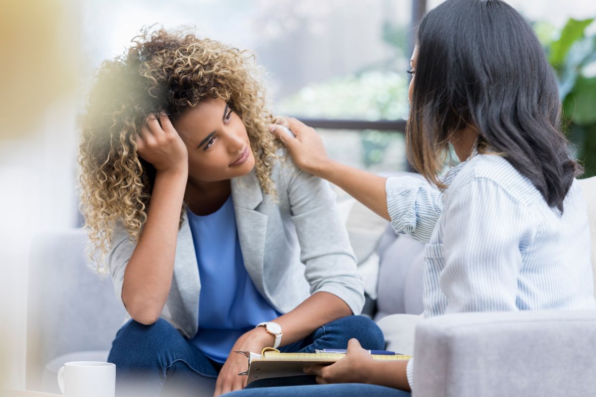 Two people are sitting on a couch. One person has their face obscured and appears to be distressed, with their head resting on their hand. The other person is gently touching the distressed person’s shoulder, seemingly offering comfort or support.