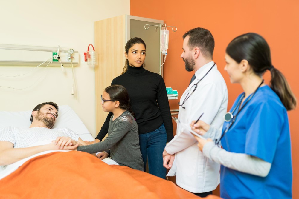 A patient lies in a hospital bed with an orange blanket. A child and an adult stand beside the bed, holding the patient's hand. Two medical professionals, one in a white coat and the other in blue scrubs, stand nearby, observing and taking notes.