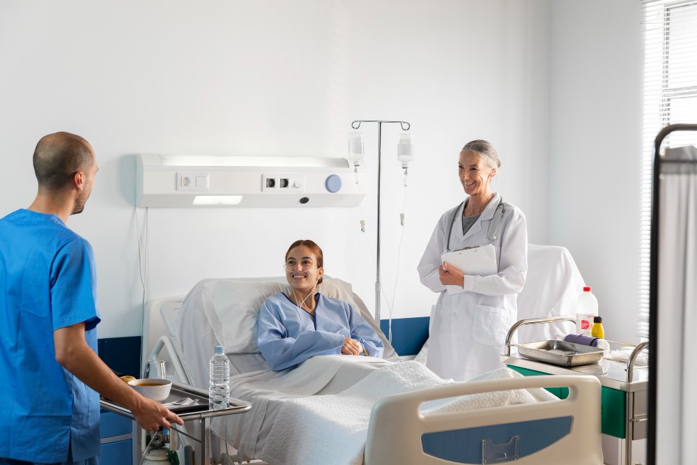 A hospital room with a patient in bed being attended by two healthcare professionals.