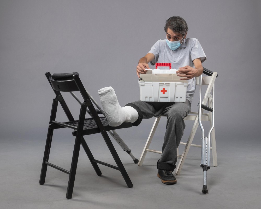 A person with a leg cast is sitting on a chair, using a walker for support, and holding a first aid kit. Another chair is positioned in front of the person to support the injured leg.