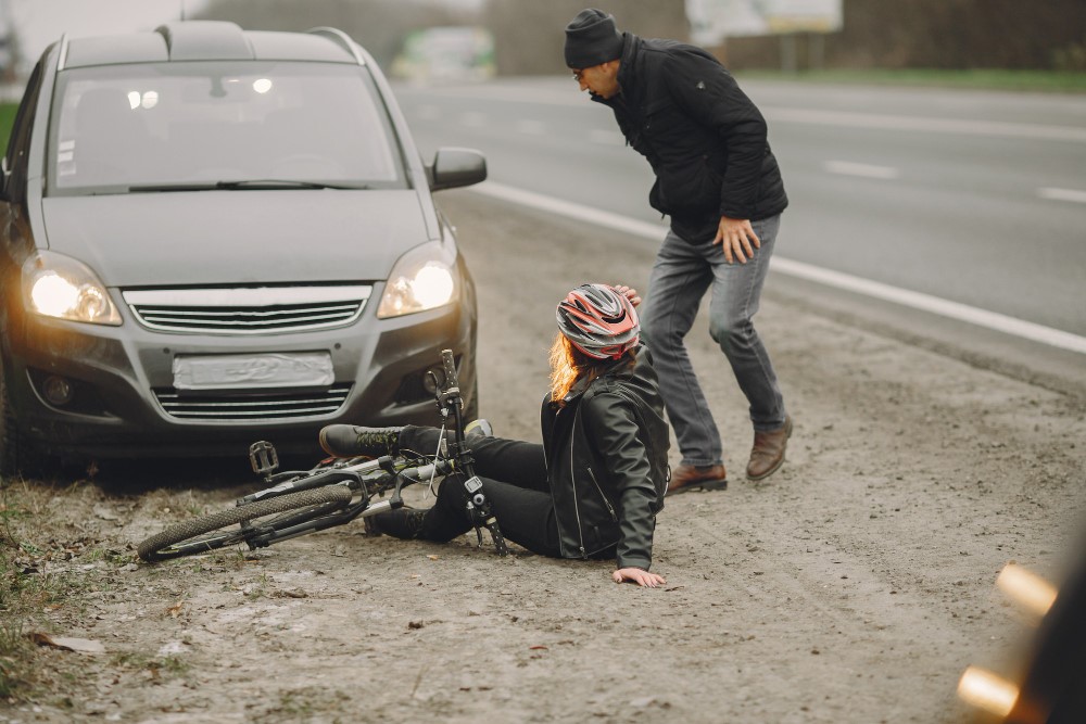 A person wearing a helmet and winter clothing is sitting on the ground next to a bicycle, appearing to have fallen. Another person is standing nearby, looking concerned. A car is parked on the side of the road next to them.