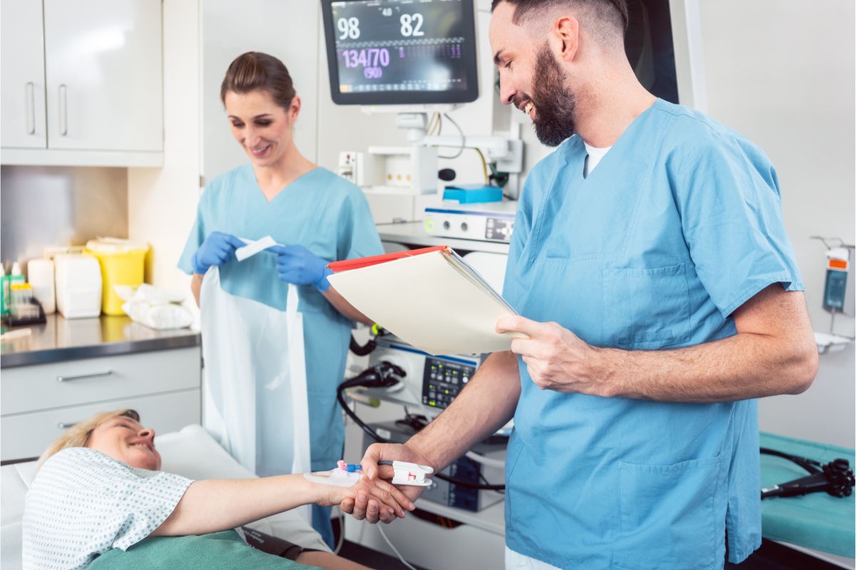 A medical team with two healthcare professionals in scrubs, smiling and interacting with a patient in a hospital room. The patient is lying in bed, receiving care, while one nurse checks their vitals on a monitor, and another shakes the patient's hand.