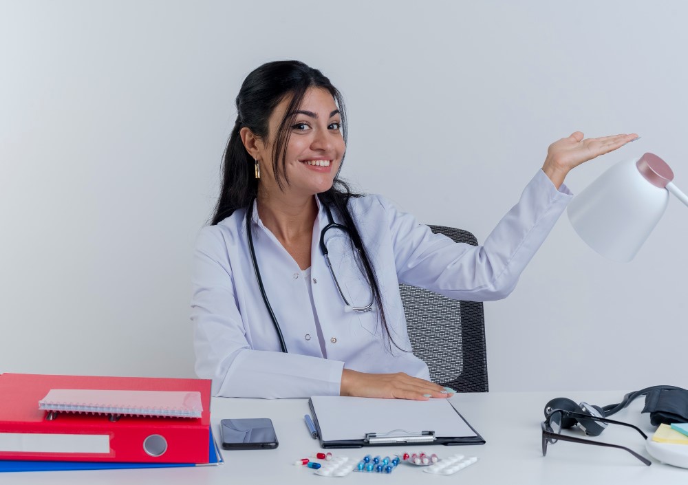 A person in a white coat, likely a doctor, is sitting at a desk with various items including a red binder, a smartphone, a clipboard, some pills, and a blood pressure monitor. The person is holding up a desk lamp with one hand.