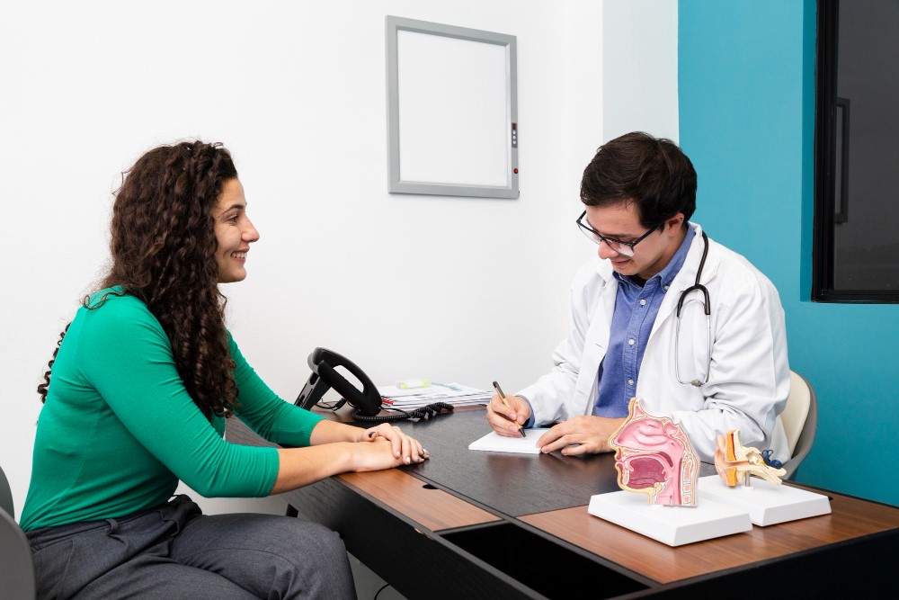 A doctor in a white coat and stethoscope is sitting at a desk, writing on a notepad, while a patient in a green sweater sits across from the doctor. On the desk are anatomical models of the human ear and other medical items.