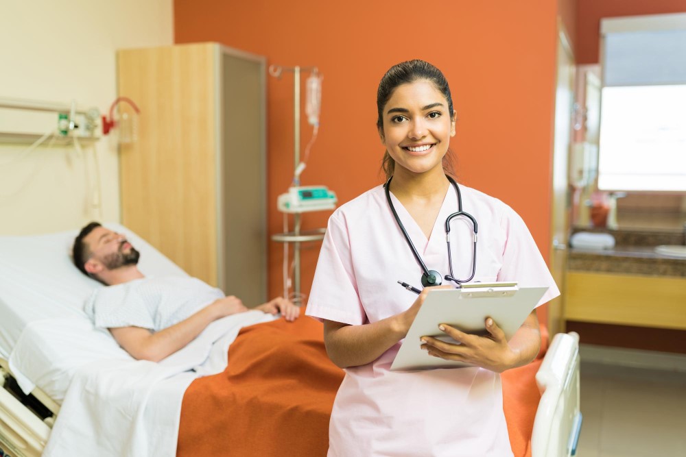 A nurse in a pink uniform holding a clipboard stands in the foreground of a hospital room. In the background, a patient lies in a hospital bed covered with an orange blanket.