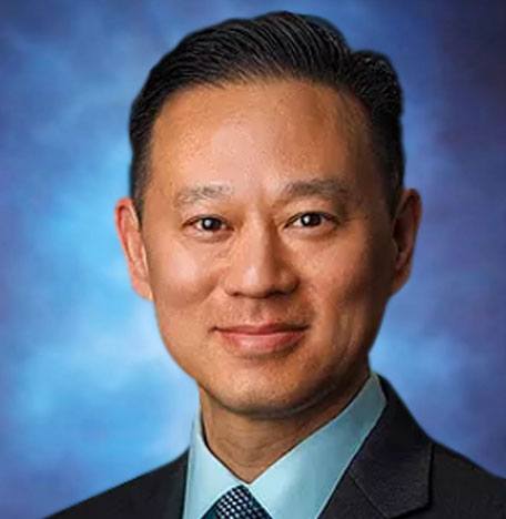 A professional headshot of a man in a suit with a calm expression, posing against a blue background. He has short dark hair and is wearing a light blue shirt with a dark blazer.