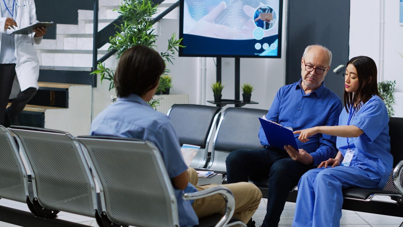 A hospital waiting area with three people seated. One person in a blue shirt is holding a clipboard, while another person in medical scrubs is pointing at it. A third person is seated with their back to the camera. A doctor is walking in the background, and a screen displaying medical information is visible.