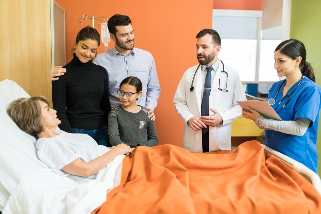 A patient lies in a hospital bed with an orange blanket, surrounded by family members and medical staff.