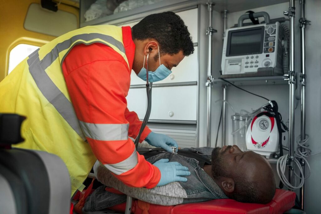 A paramedic in a yellow and red uniform is using a stethoscope to check the chest of a patient lying on a stretcher inside an ambulance. Medical equipment is visible in the background.