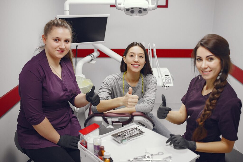 Three individuals in a dental clinic, two wearing purple uniforms and black gloves, and one in casual clothing, all giving thumbs up. Dental equipment and tools are visible in the background.