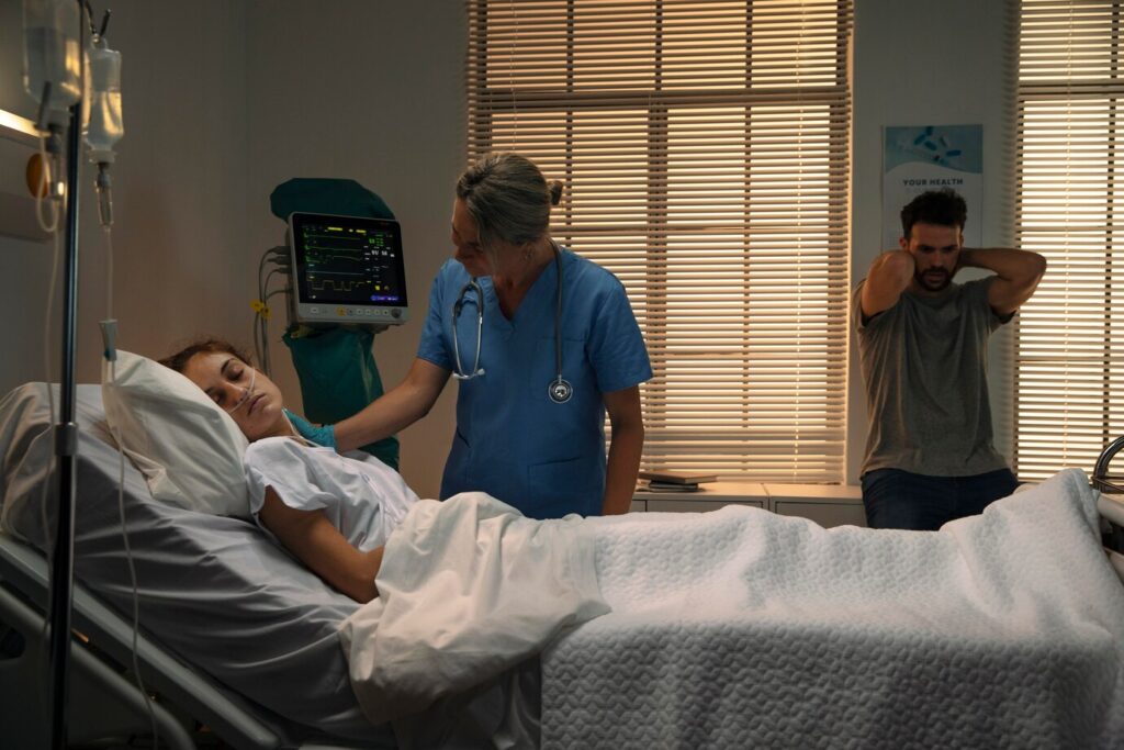 A medical professional in blue scrubs is attending to a patient lying in a hospital bed, with a person standing in the background.