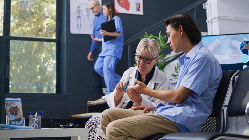 A healthcare professional in a white coat is showing a medical device to a seated individual in a blue shirt. In the background, another healthcare worker in blue scrubs is walking down the stairs, while a person in a blue shirt is standing nearby. The setting appears to be a medical facility with anatomical posters on the wall and a large window letting in natural light.