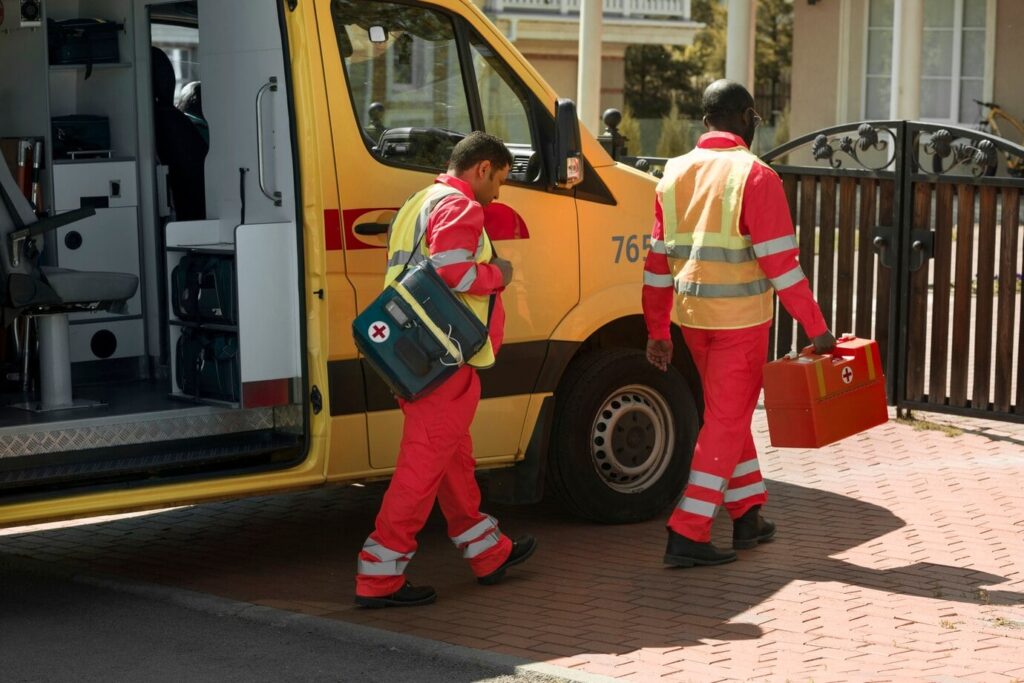 Two emergency medical responders in red uniforms and reflective vests are walking away from a yellow ambulance. One is carrying a green medical bag with a red cross, and the other is holding an orange medical kit with a red cross. The back door of the ambulance is open, revealing the interior with medical equipment.