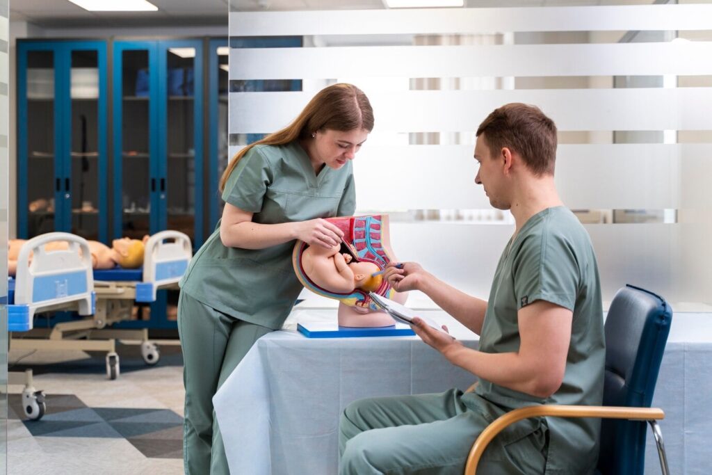 Two medical professionals in green scrubs are examining a model of a pregnant woman's anatomy, focusing on the fetus inside the womb. They are in a medical training room with hospital beds and medical mannequins in the background.