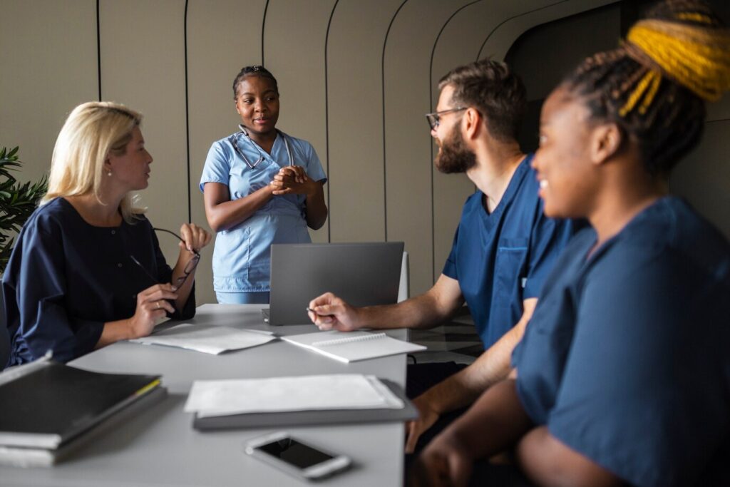 A group of healthcare professionals in blue scrubs having a meeting around a table with notebooks, a laptop, and a smartphone.