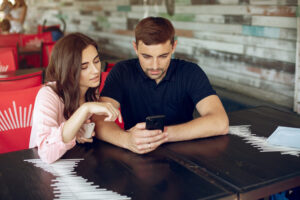 A young woman in a pink shirt and a young man in a black polo sitting at a restaurant table, looking intently at a black smartphone screen.