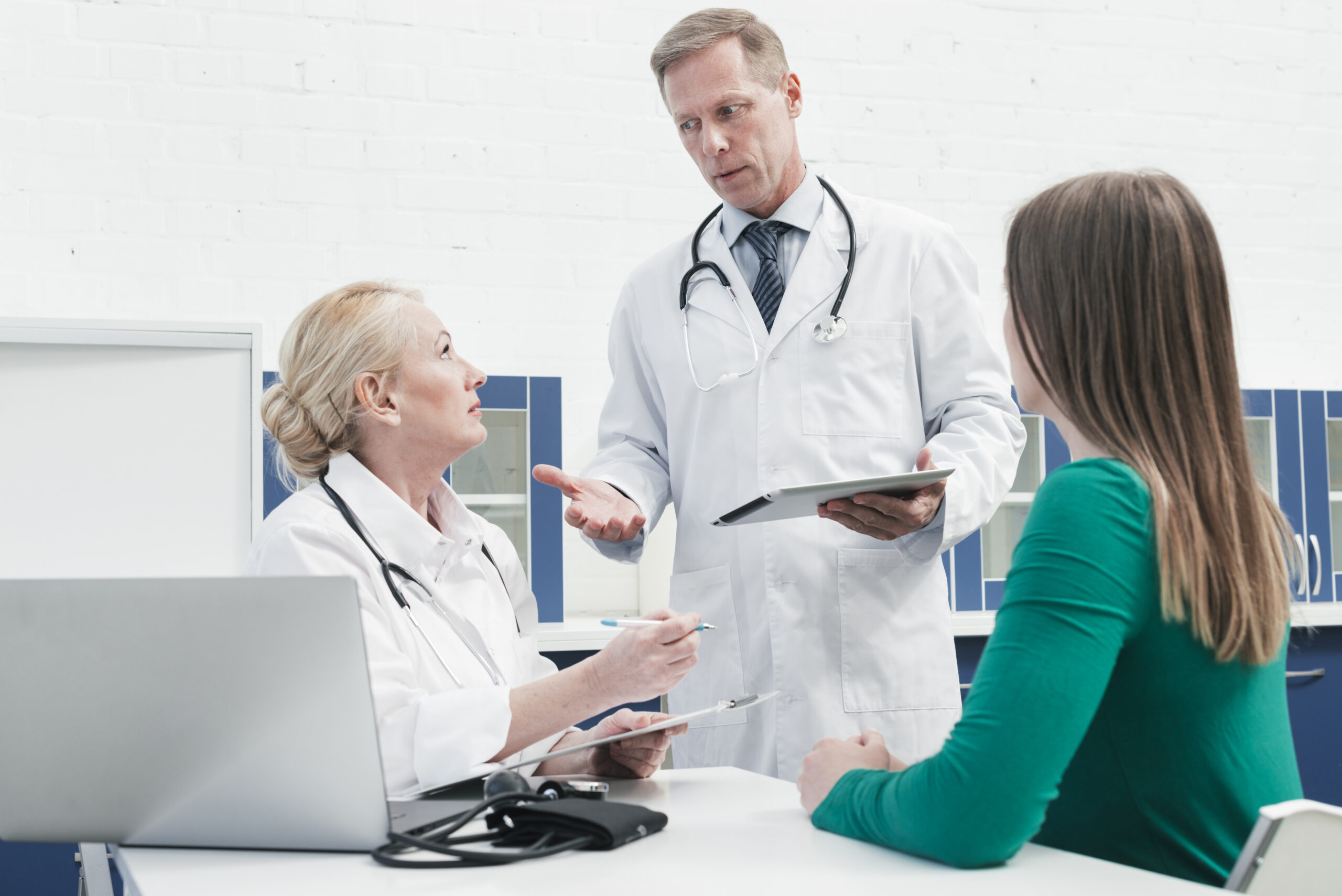 A male and female doctor in white coats discussing medical results on a tablet and clipboard with a patient at an urgent care clinic.