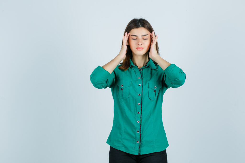Young woman with closed eyes holding both temples, showing signs of headache, stress, fatigue, or migraine against a plain background.