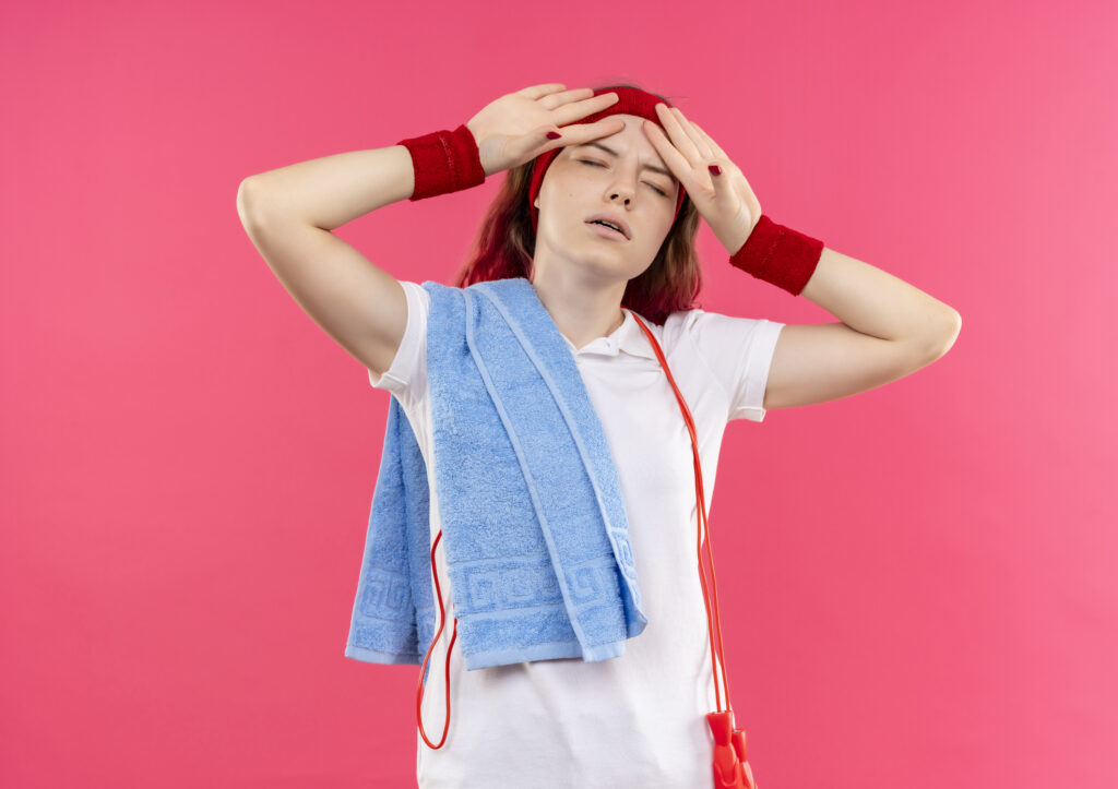 Woman experiencing heat exhaustion and fatigue after exercise while holding her forehead with a towel over her shoulder.