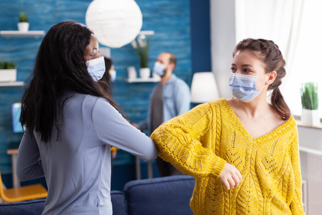 Two masked patients greeting each other with an elbow bump in a modern medical clinic while practicing social distancing.