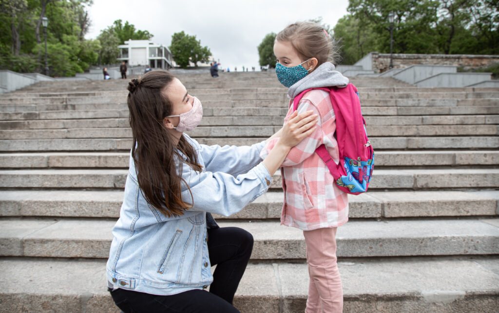 Mother helping her young daughter with her backpack while both wear face masks on school steps during the COVID-19 pandemic.