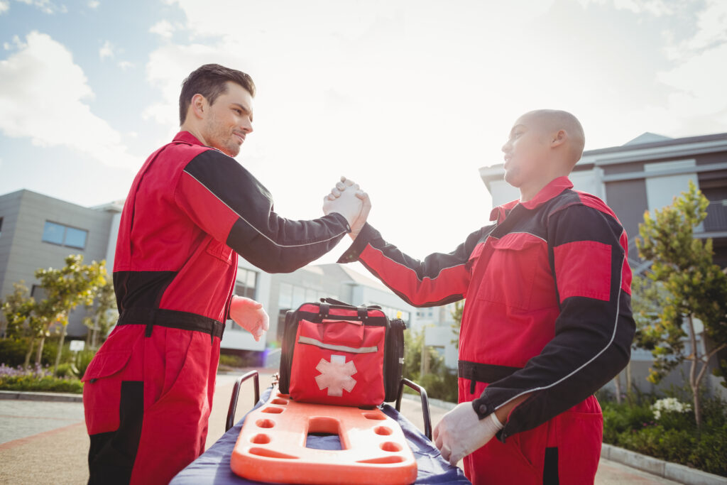 Two emergency medical responders shaking hands beside a stretcher and medical bag outside an urgent care facility.