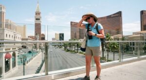 A weary female tourist wipes sweat from her brow while standing on an elevated pedestrian bridge over the Las Vegas Strip, holding a water bottle with iconic casinos like The Venetian and Wynn in the background.
