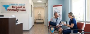 A medical professional shows an education app on a tablet to a male patient with a left arm sling and blue knee tape in the waiting room of Sahara West Urgent Care & Primary Care. The 'SAHARA WEST' logo is on the wall. A guide poster reads 'LAS VEGAS Acute Rehab and Our Care'. An X-ray room is visible in the hallway.
